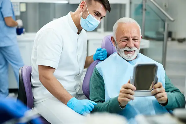 Happy, senior patient looking at himself in a mirror with dental implants at Periodontal Surgical Arts in Austin, TX Happy, senior patient looking at himself in a mirror with dental implants at Periodontal Surgical Arts in Austin, TX